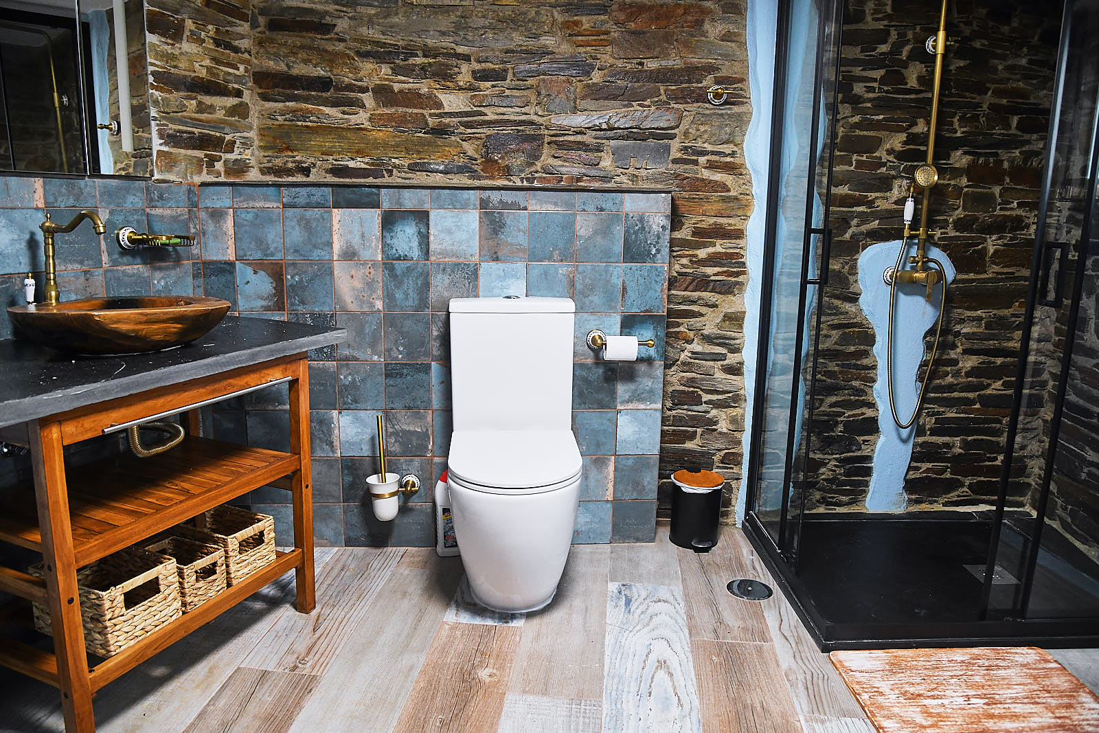 Bathroom with wooden vanity and carved stone basin, brass wall faucet, toilet and black-framed walk-in shower against an exposed schist wall